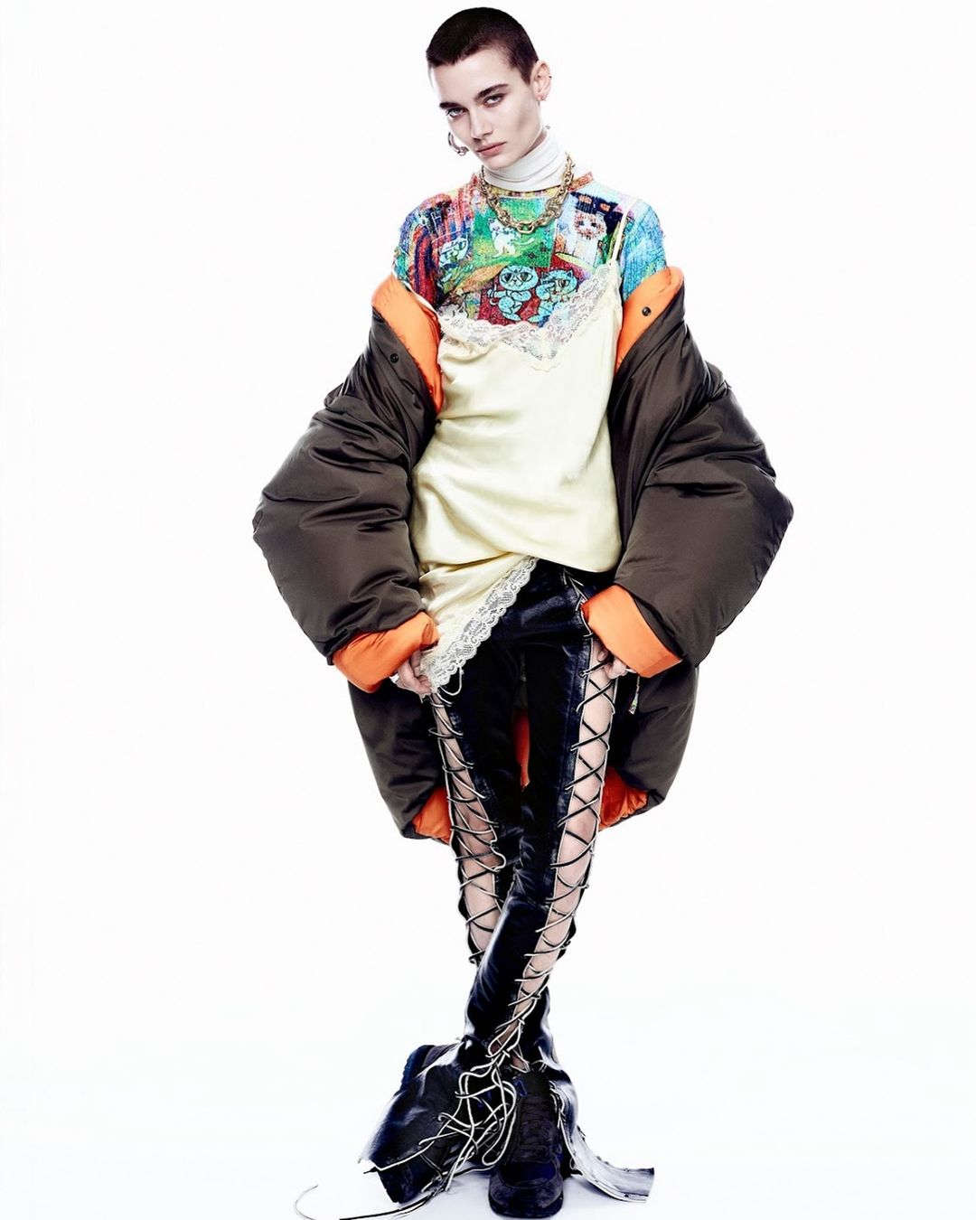 Fashionable woman with a buzzcut poses in a colorful patterned top, lace-trimmed dress, oversized puffer jacket, and lace-up boots in a photo studio.