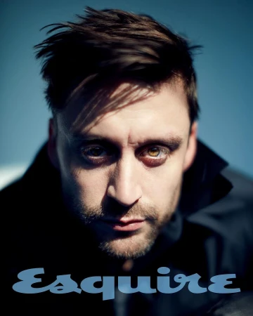Portrait of adult male with dark hair and light eyes, wearing a black collared shirt with Esquire magazine logo in the foreground.