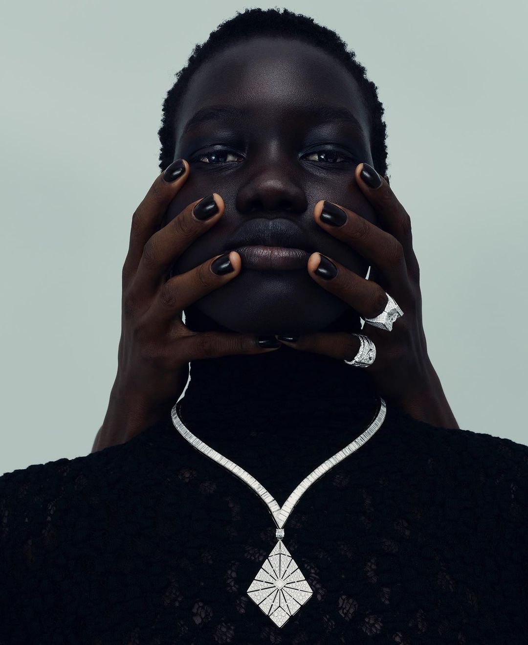 Close-up of a Black woman with dark hair and black nail polish wearing a diamond necklace, posing with hands framing her face.