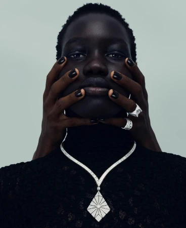 Close-up of a Black woman with dark hair and black nail polish wearing a diamond necklace, posing with hands framing her face.