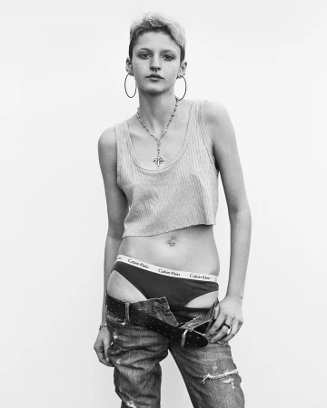 Monochrome portrait of a model with short hair wearing a ribbed crop top, hoop earrings, and unbuttoned jeans against a plain backdrop.