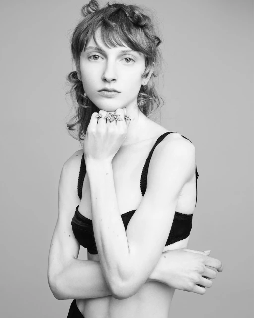 Black-and-white portrait of a young adult female model wearing a black bra and multiple rings, posing against a neutral background.