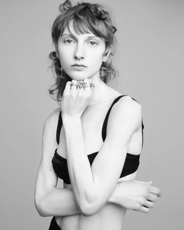 Black-and-white portrait of a young adult female model wearing a black bra and multiple rings, posing against a neutral background.
