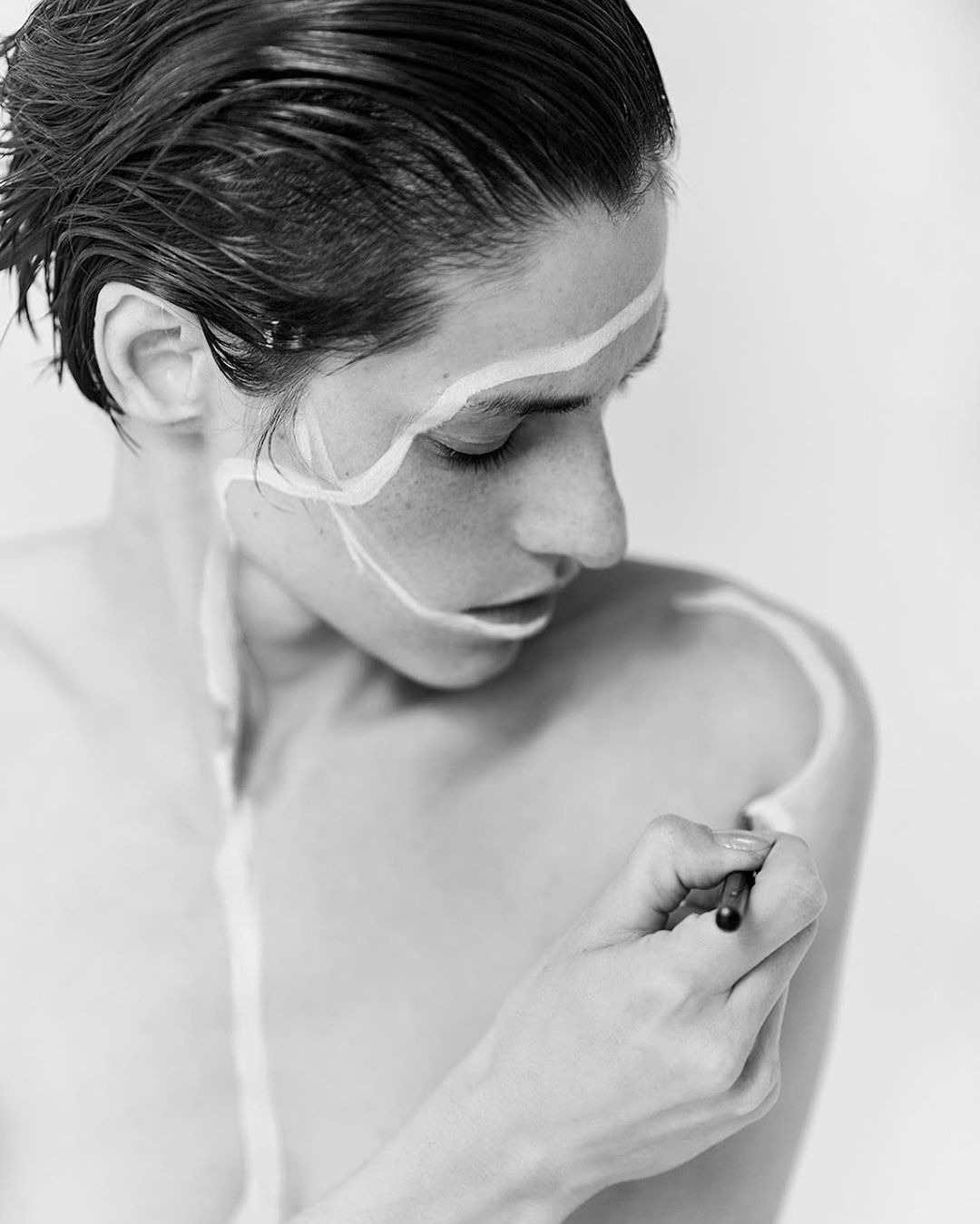 Monochrome close-up of a shirtless adult with paint on their face and body, seen indoors in a studio setting, holding a paintbrush.