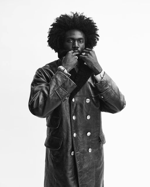 A Black male with an afro wearing a leather trench coat in a studio setting, creating a high-contrast black and white fashion portrait.