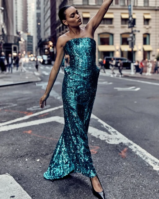 Woman in a blue sequin gown strikes a pose on a city street with pedestrians and buildings in the background during daytime.