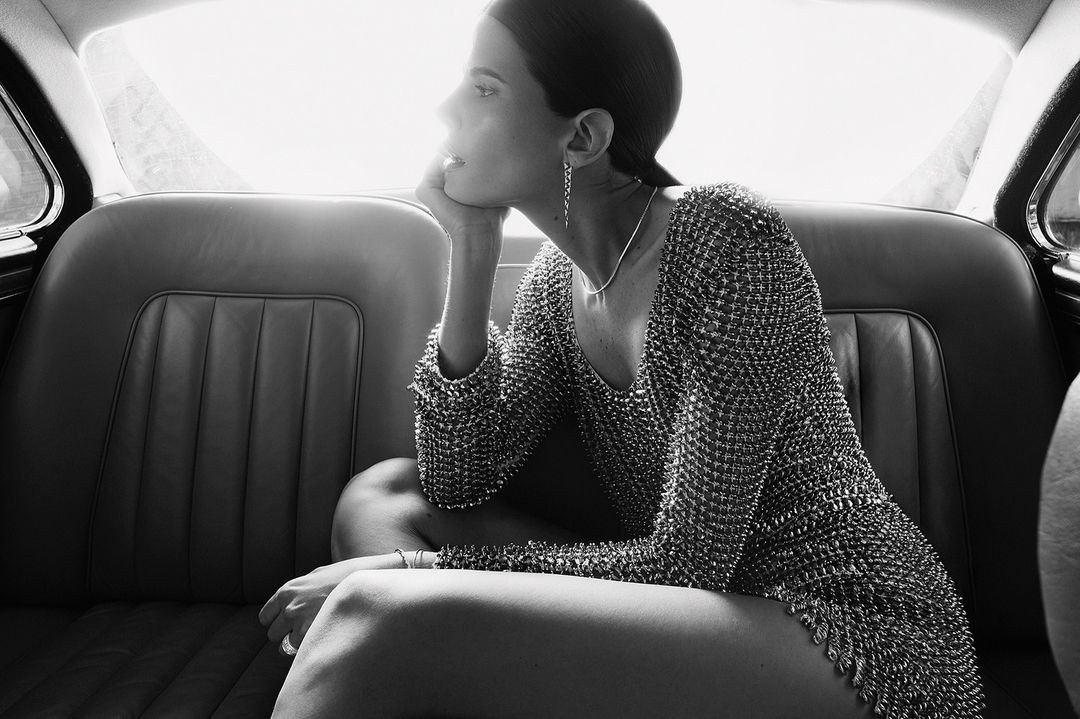 Woman in metallic dress sits in a classic car, black and white. Elegant indoor shot with soft light and thoughtful expression.
