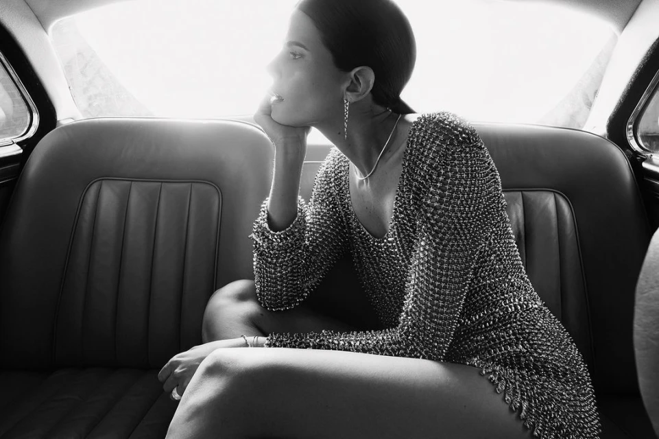 Woman in metallic dress sits in a classic car, black and white. Elegant indoor shot with soft light and thoughtful expression.