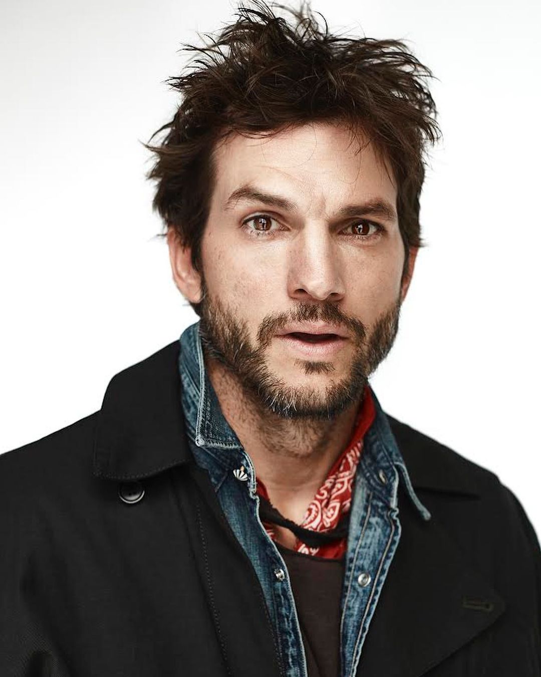 Close-up portrait of a handsome adult male with a beard wearing a blue denim jacket and a black coat against a white backdrop.
