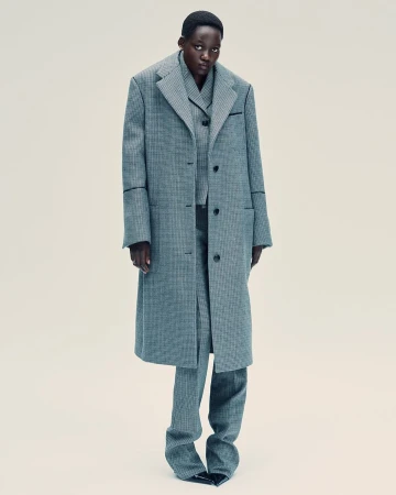 Fashionable woman poses in a patterned overcoat and matching suit, complemented by black pointed shoes against a neutral backdrop.