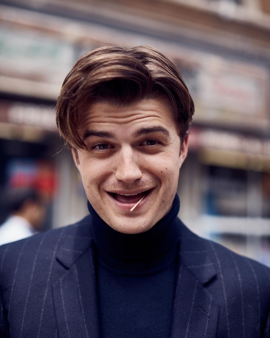 Handsome man in a pinstripe suit smiles with a toothpick in his mouth, portrait shot in front of an urban storefront.