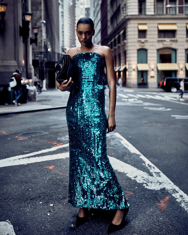 Elegant woman in a blue sequin gown, holding a clutch, poses in a city street with tall buildings in the background. Fashion editorial shot.