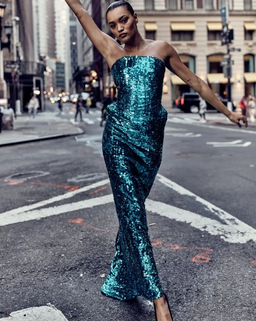 Adult female in a blue sequin gown posing on a city street with buildings and pedestrians in the background, full-body shot.