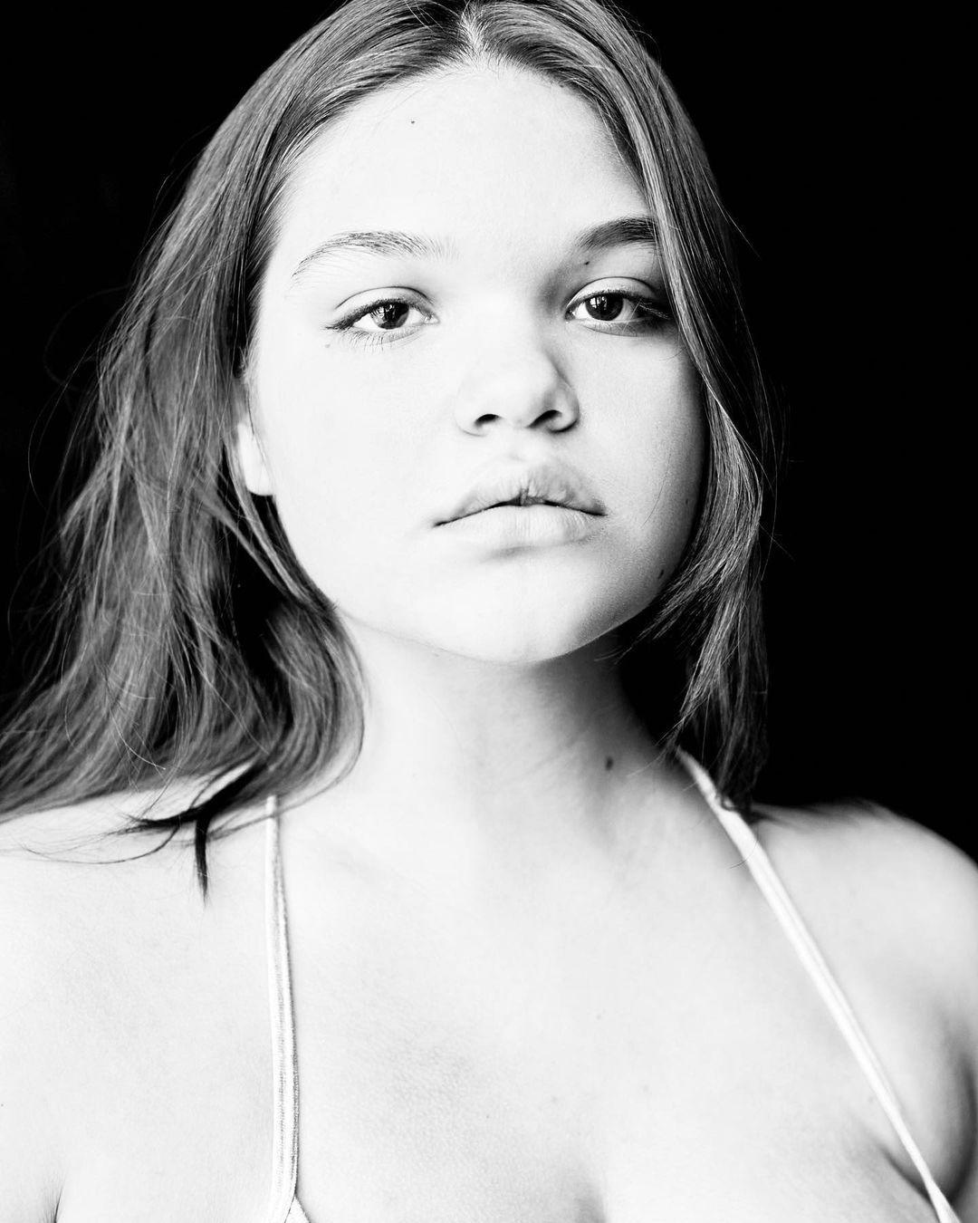 Monochromatic portrait of a young woman with long hair, gazing directly at the camera against a plain black backdrop in a close-up shot.