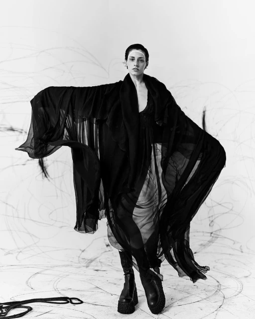 A person wearing a long, flowing, black gown stands in a room with dark scribbles on the walls and floor and wears heavy, black boots.