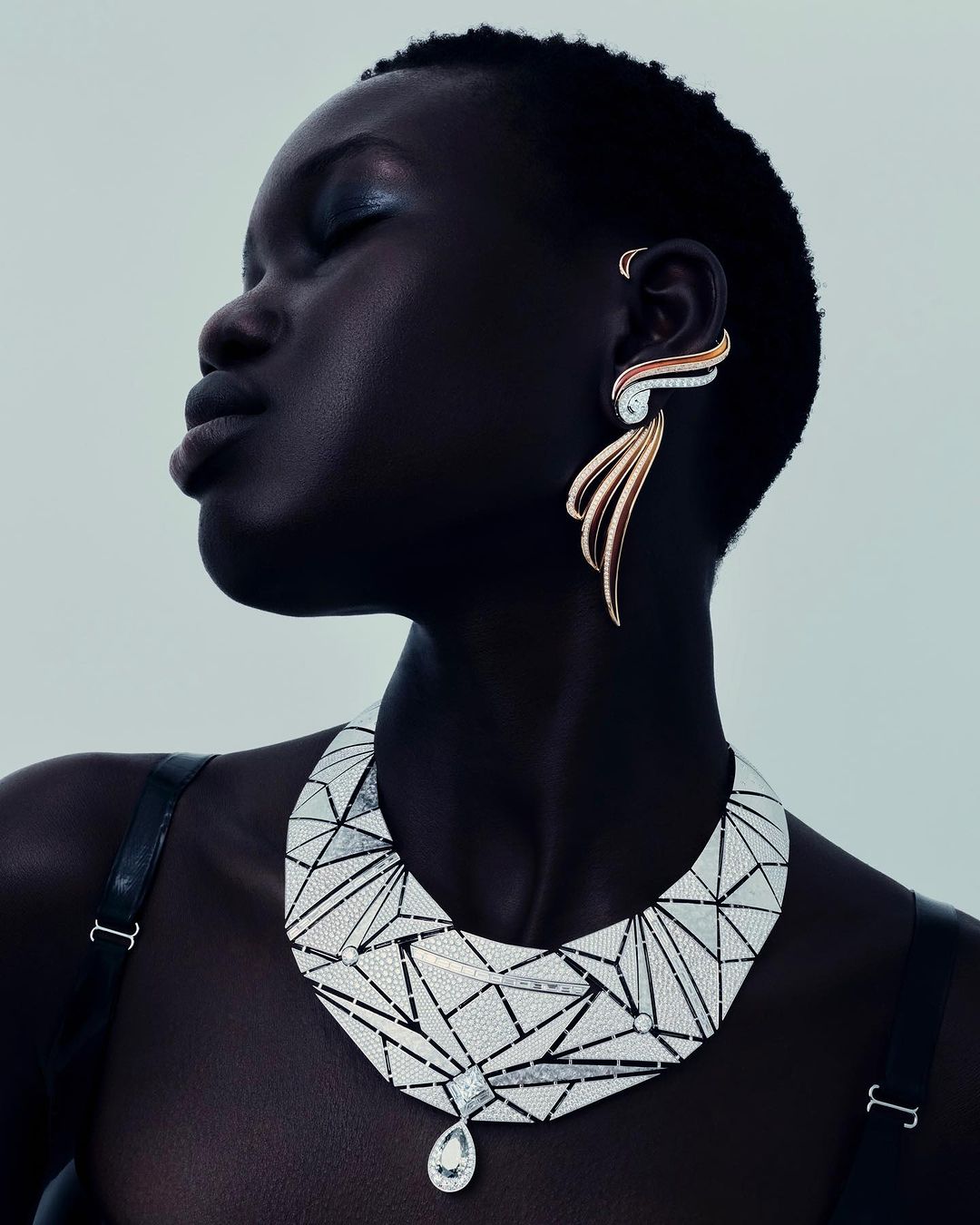 Close-up of a woman with dark skin wearing sculptural earrings and a geometric necklace against a neutral background in studio lighting.