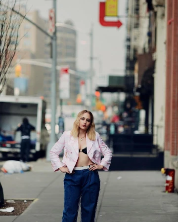Woman with blonde hair wearing a pink jacket and blue trousers standing on a city sidewalk