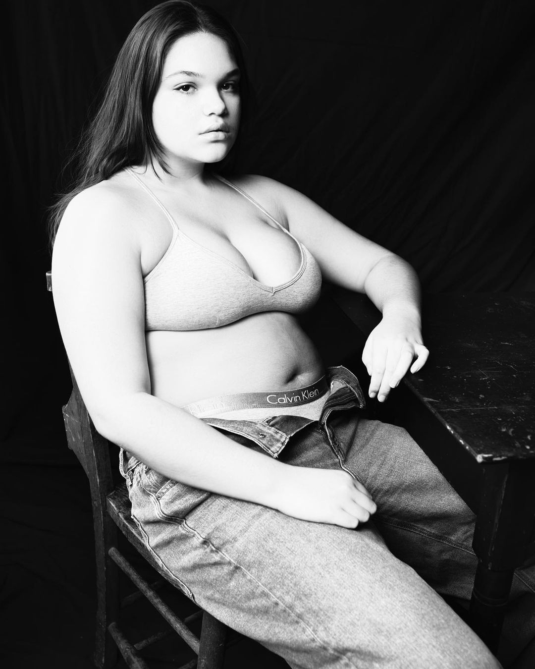Black and white portrait of a woman in a bra and jeans sitting at a table with a dark background, looking at the camera.