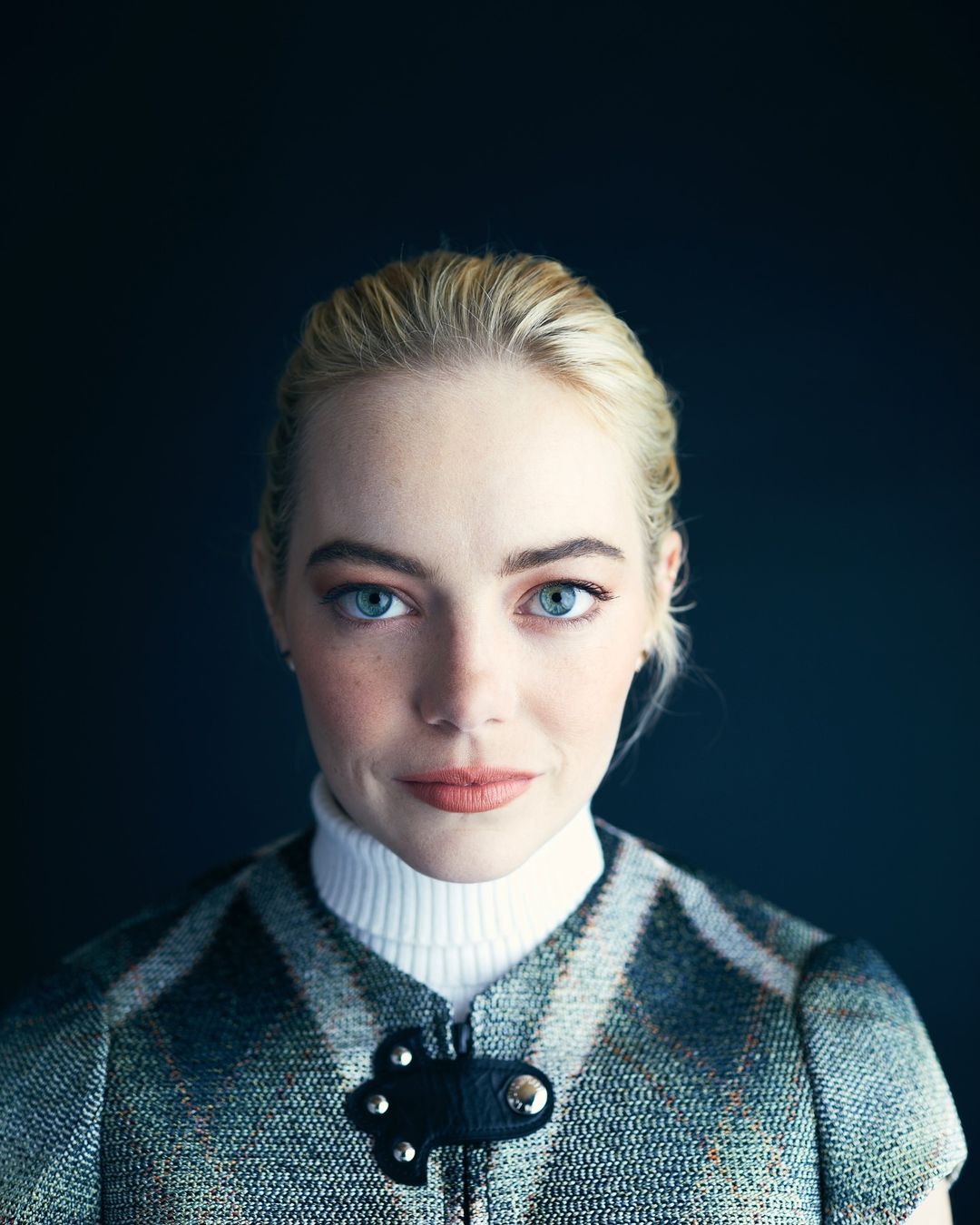 Close-up portrait of a fair-skinned adult female with blonde hair and blue eyes, wearing a tweed vest over a white turtleneck against a dark background.
