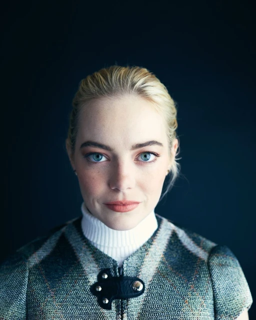 Close-up portrait of a fair-skinned adult female with blonde hair and blue eyes, wearing a tweed vest over a white turtleneck against a dark background.