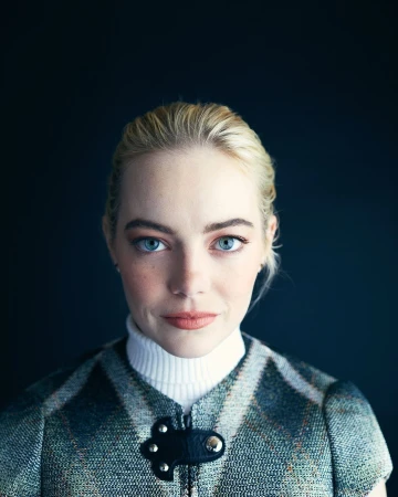 Close-up portrait of a fair-skinned adult female with blonde hair and blue eyes, wearing a tweed vest over a white turtleneck against a dark background.