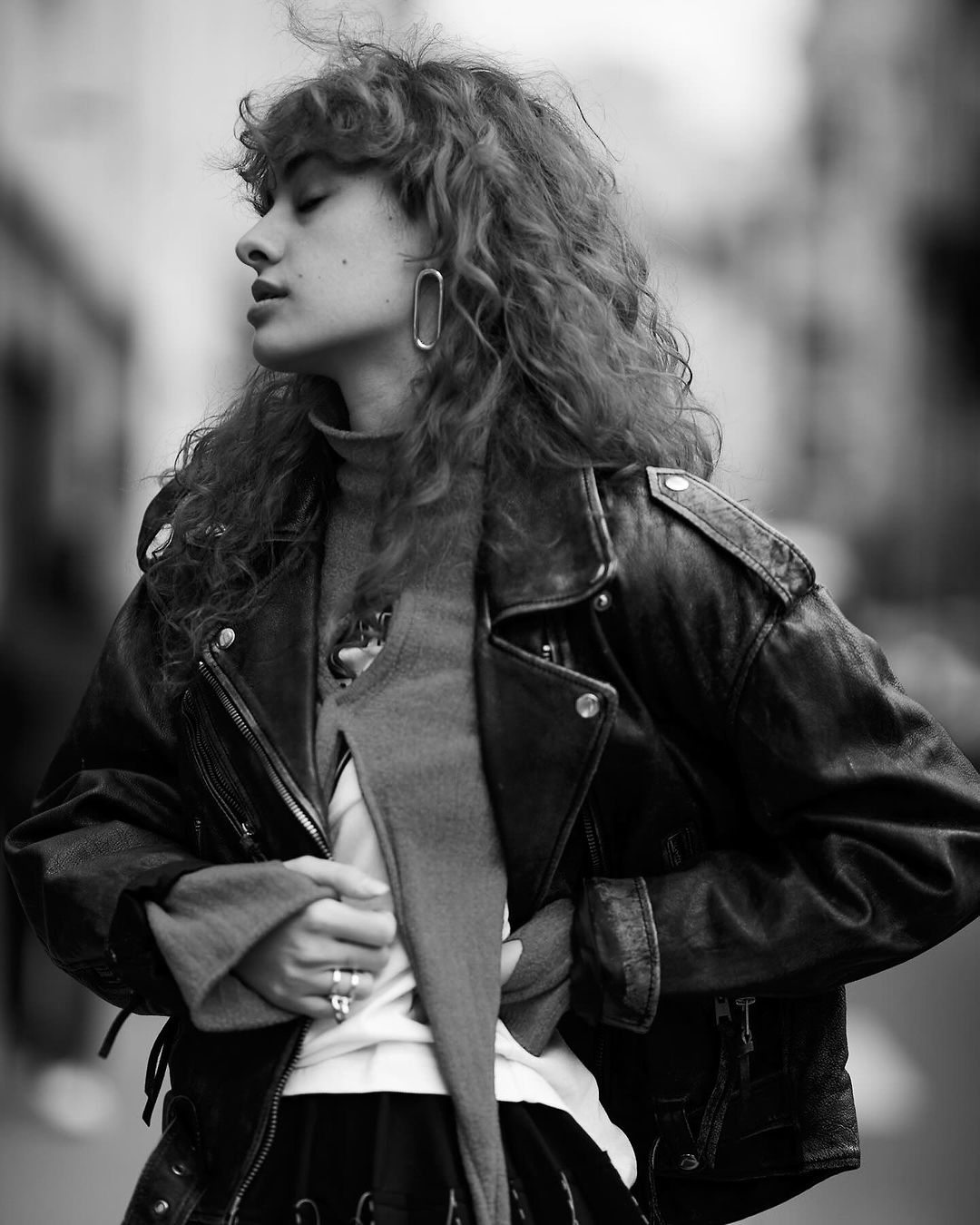 Monochrome portrait of a young woman with curly hair wearing a leather jacket, a turtle neck sweater and earrings in a street setting.