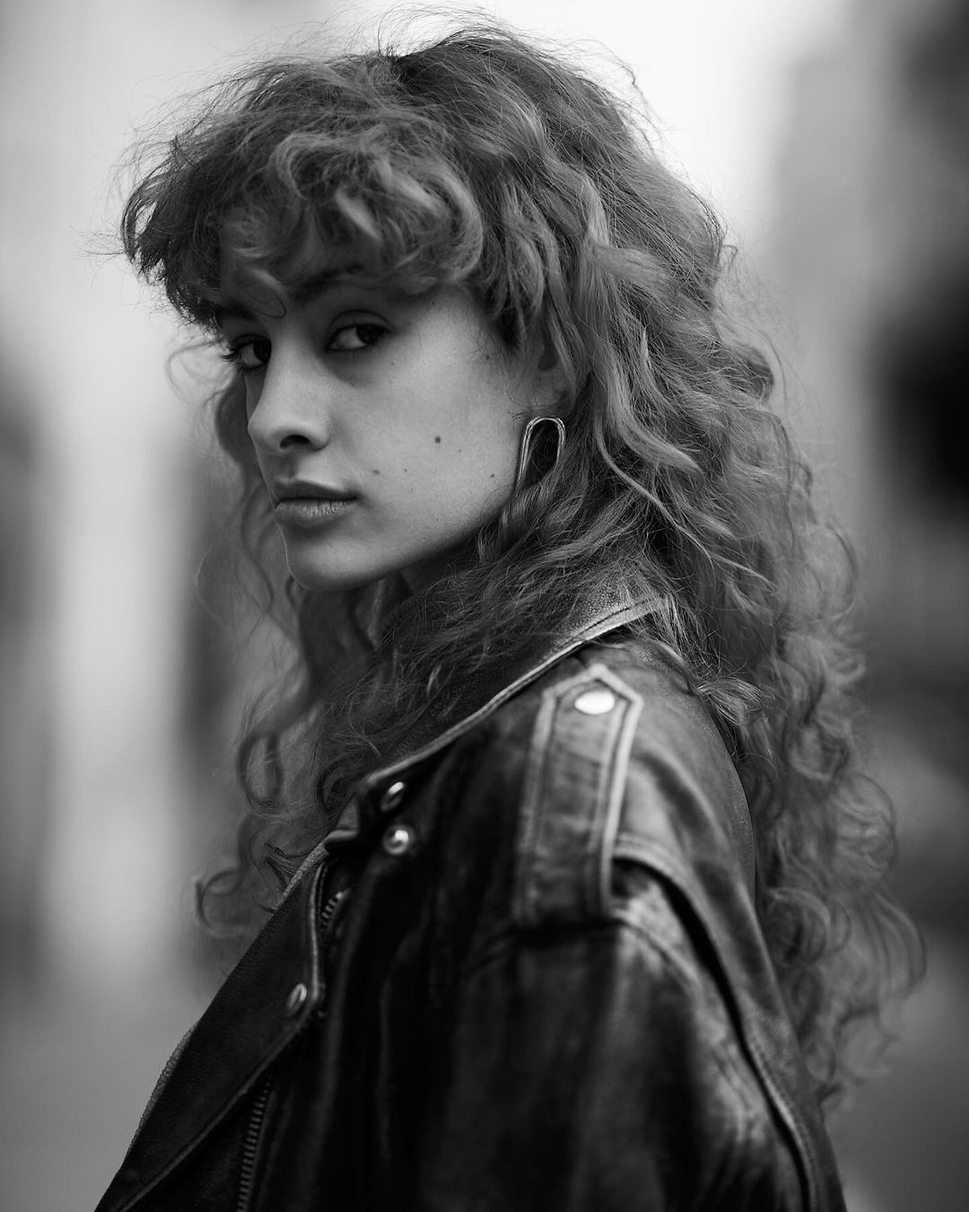Black and white portrait of a woman with curly hair, wearing a leather jacket, looking over her shoulder in an outdoor urban setting.