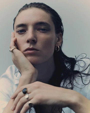 Close-up portrait of a woman with wet hair resting her chin on her hand, wearing rings and hoop earrings, against a light background.