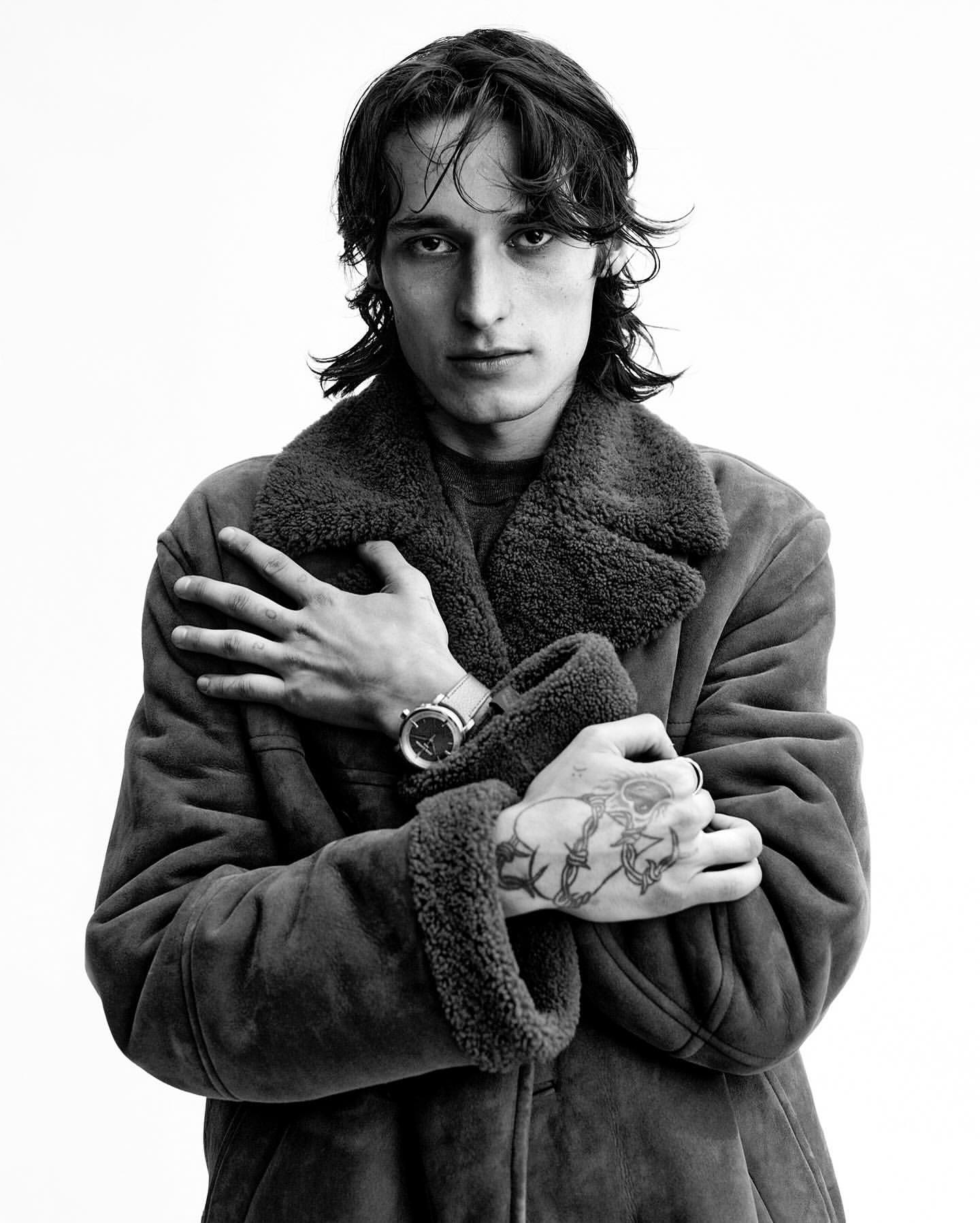 Black and white portrait of a young man in a shearling coat with arms crossed, showcasing hand tattoos and a wristwatch against a plain background.