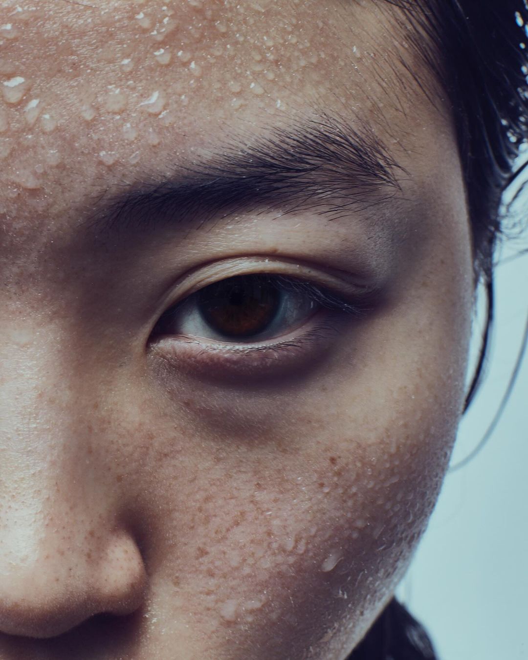 Close-up studio shot of an adult with dark hair and freckled skin, drops of moisture on forehead, eye-level view, cool tones.