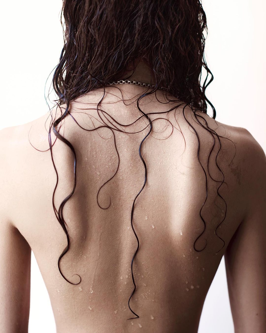 Close-up shot of a woman's bare back with wet, dark, wavy hair cascading down; skin glistening with water droplets against a bright backdrop.