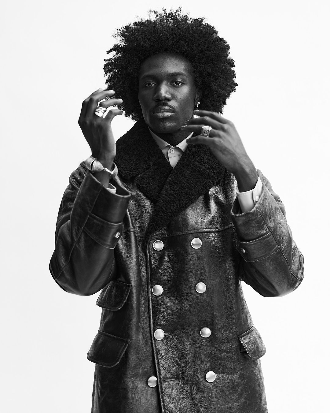 Headshot of a Black male adult wearing a black, double-breasted leather coat with a shearling collar against a neutral backdrop.