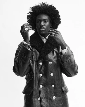 Headshot of a Black male adult wearing a black, double-breasted leather coat with a shearling collar against a neutral backdrop.