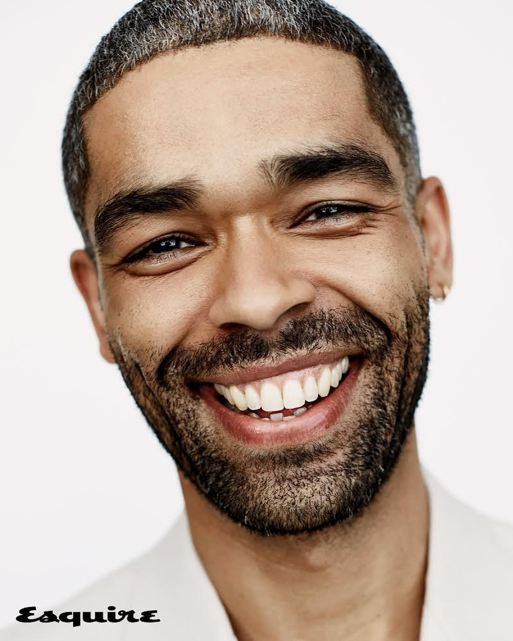 Close-up portrait of a smiling black man with short dark hair and facial stubble against a white background.