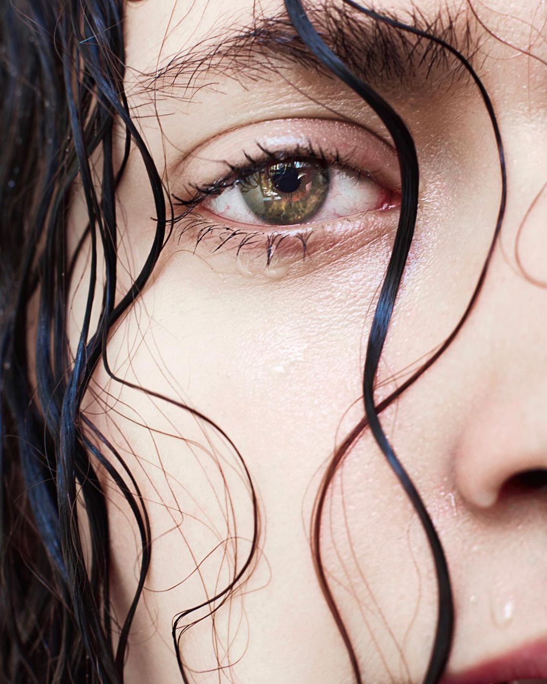 Close-up of a woman's face with a tear rolling down her cheek. The image shows the woman's eye, eyelashes, and wet hair.