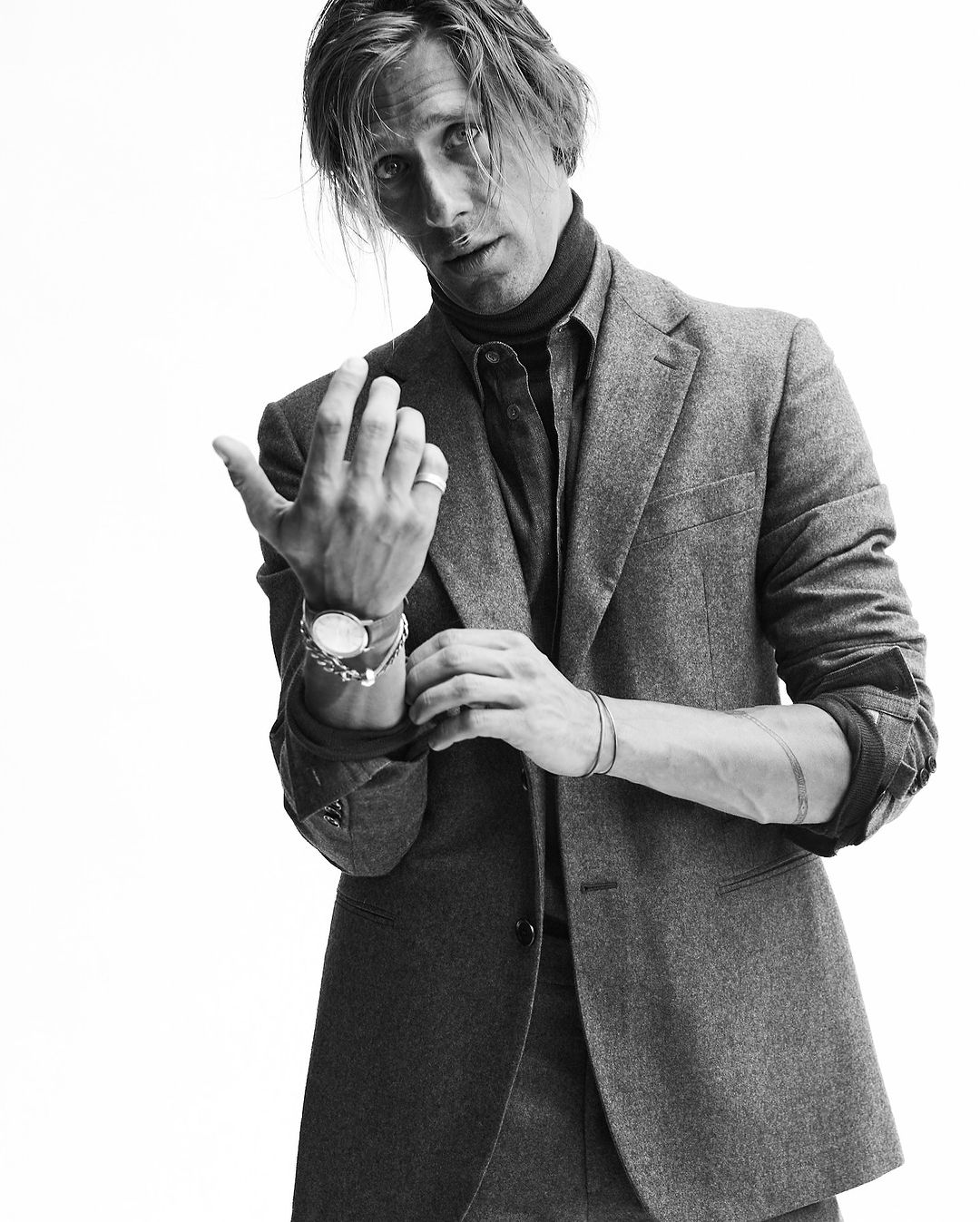 Man wearing a grey tweed blazer and dark turtleneck adjusting his sleeve against a white background
