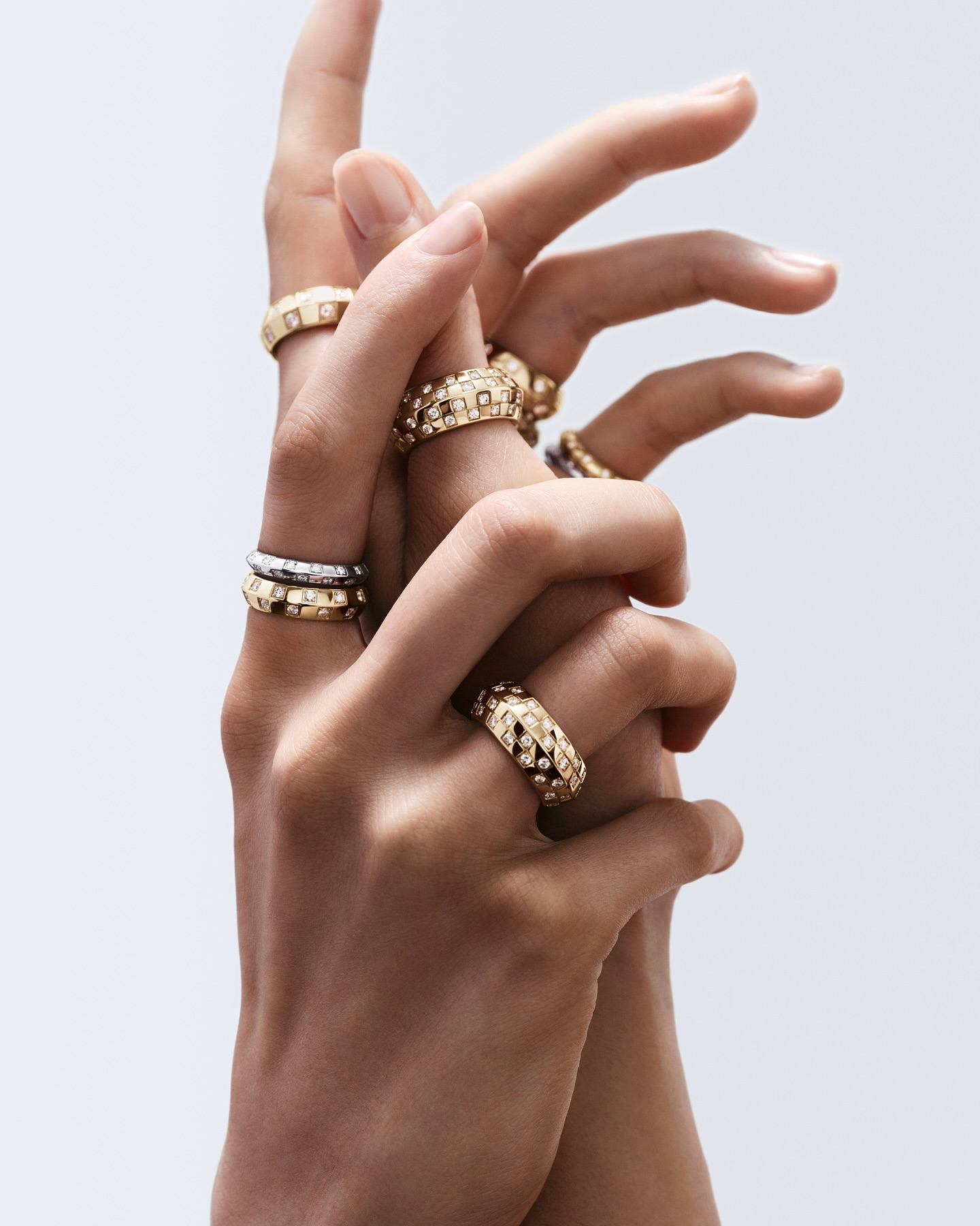 Close-up of two hands interlocked wearing gold rings with diamond details on a light grey background for a jewelry advertisement.