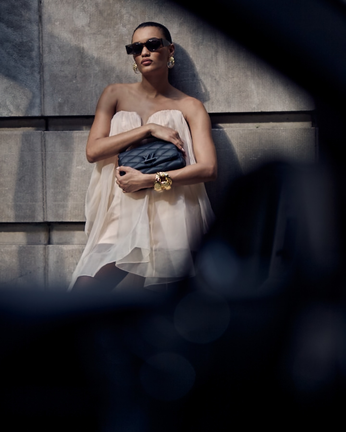 Woman wearing a dress and sunglasses poses with a handbag in front of a concrete wall, accessorized with gold jewelry and a dark handbag.