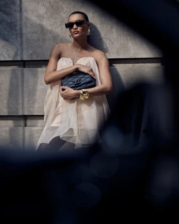 Woman wearing a dress and sunglasses poses with a handbag in front of a concrete wall, accessorized with gold jewelry and a dark handbag.