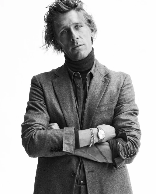Black and white portrait of a man in a suit with arms crossed, wearing a turtleneck, dress shirt, and wristwatch against a white background.
