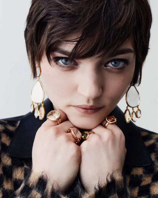 Close-up studio portrait of a woman wearing oversized stone earrings and gold rings with gems on her fingers, set against a neutral background.