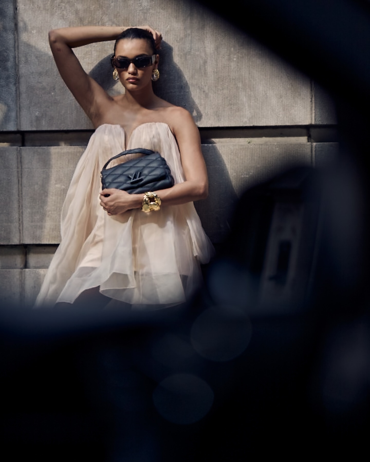 Model in strapless tulle dress wearing sunglasses and holding a quilted handbag against a stone wall