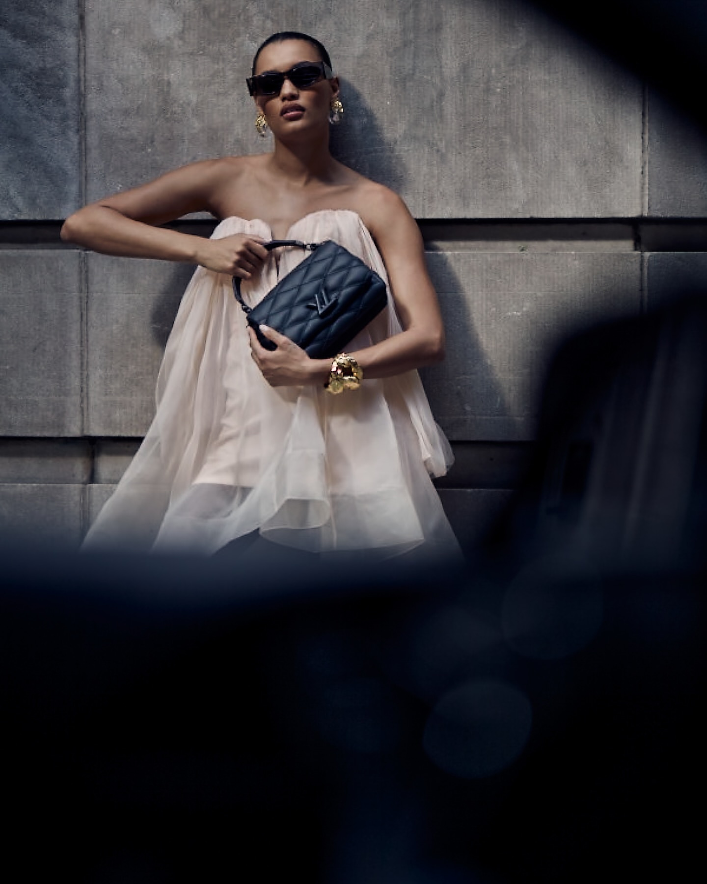 Elegant woman poses with black quilted purse and gold jewelry against a textured grey wall, wearing sunglasses and a draped peach dress.