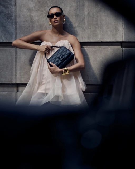 Elegant woman poses with black quilted purse and gold jewelry against a textured grey wall, wearing sunglasses and a draped peach dress.