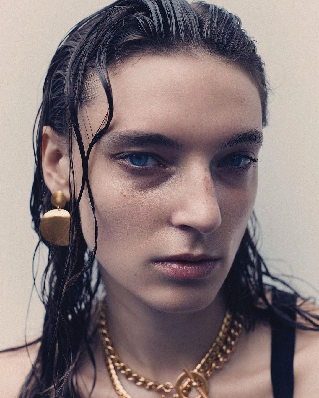 Close-up portrait of a young fair-skinned woman with wet dark hair, blue eyes, gold chain necklace, and matching earrings, indoors.