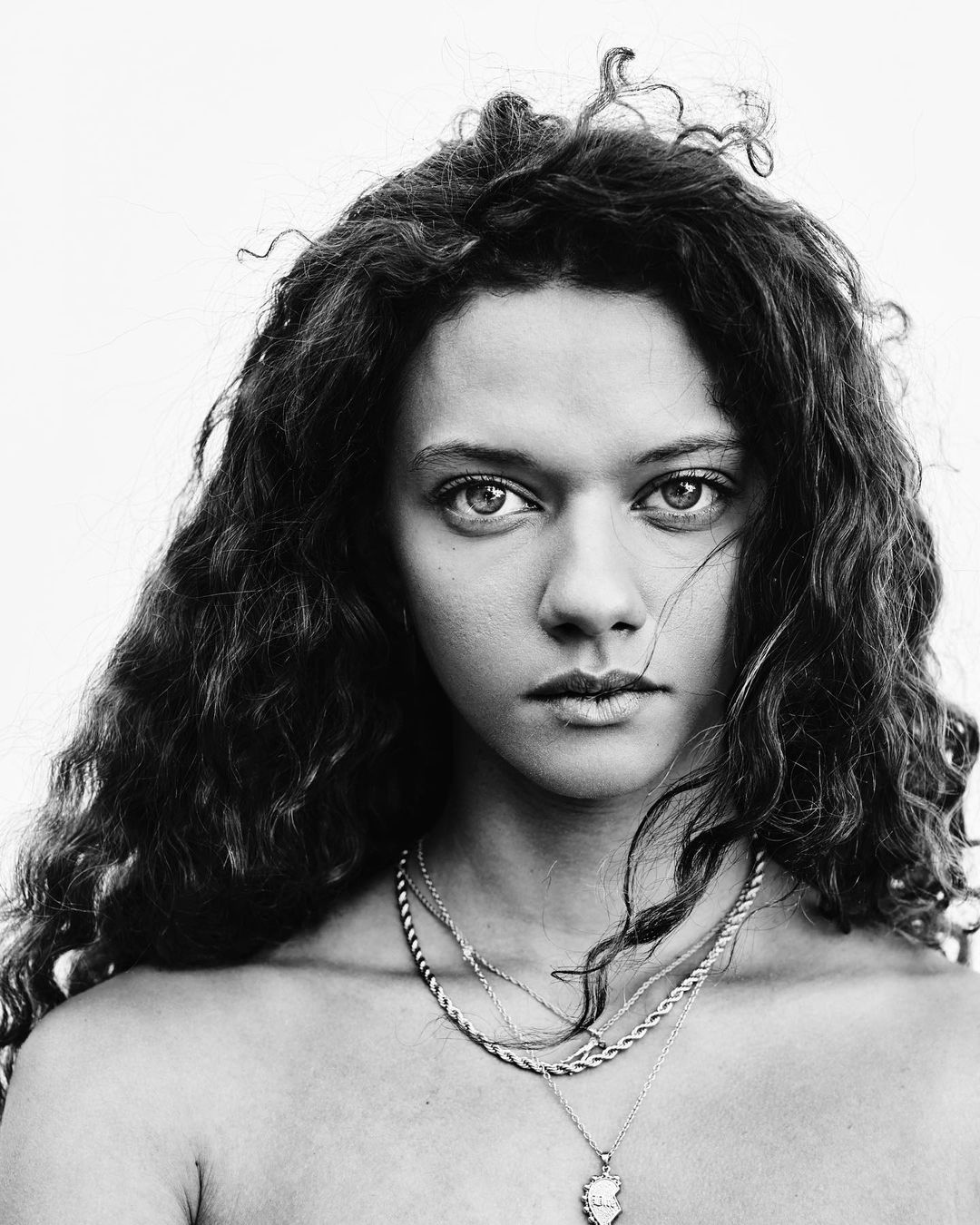 Striking black and white portrait of a young adult female with curly hair, silver necklaces, and direct eye contact against a plain white backdrop.
