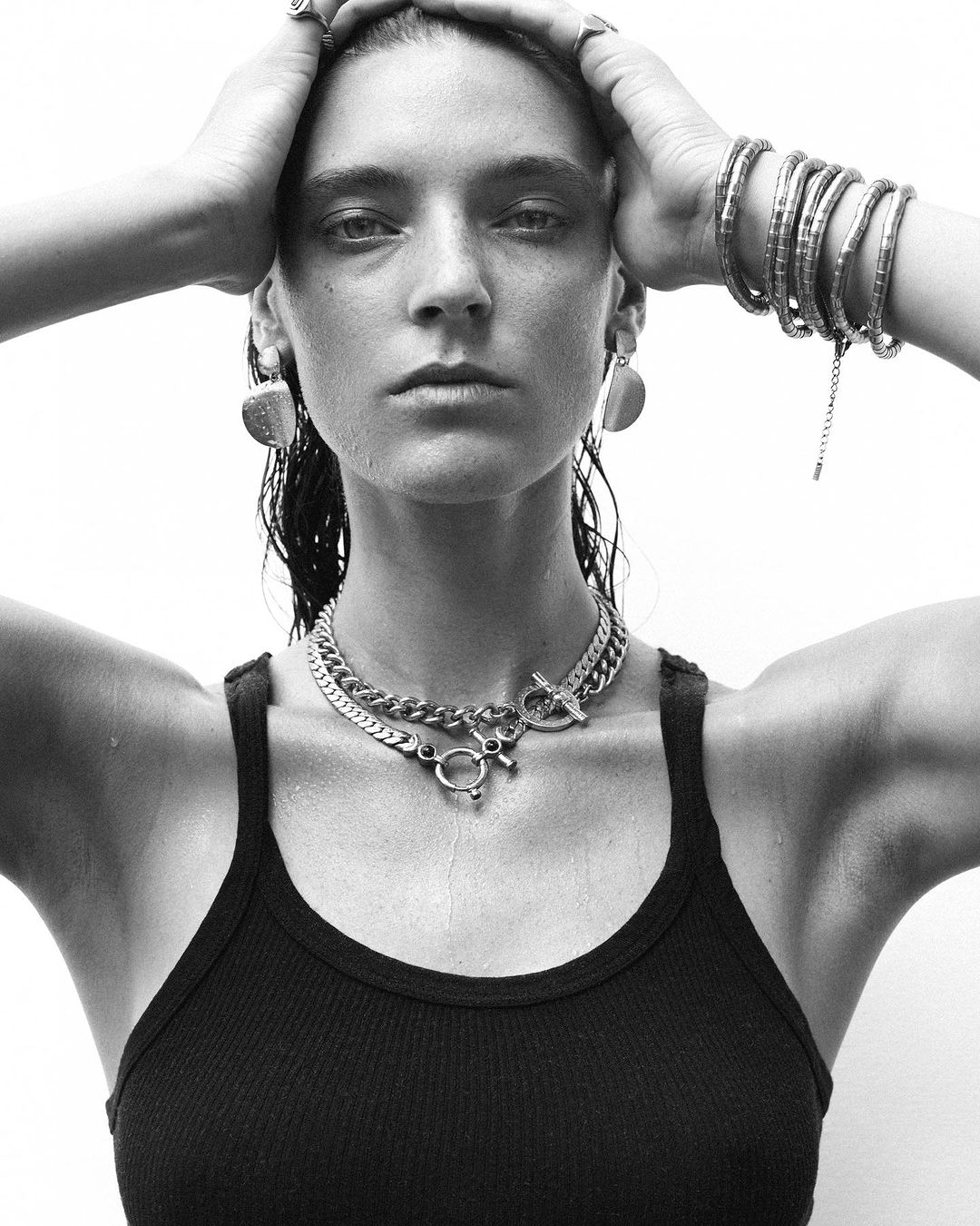 Monochrome portrait of a woman wearing layered silver necklaces, bracelets, and rings against a plain white background.