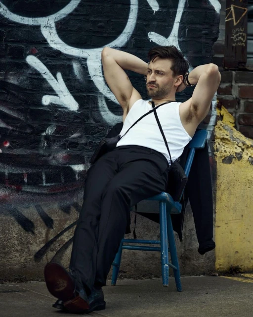 Man in a white tank top and black pants sits on a blue chair against a graffiti wall in an urban setting, looking away with a relaxed posture.
