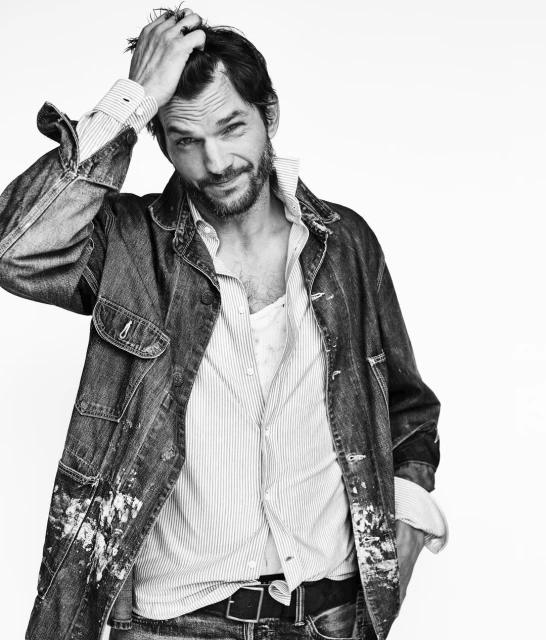 Portrait of a relaxed adult male with a beard, wearing a denim jacket over a button-down shirt, posing against a white backdrop.