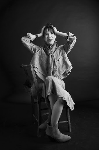 Monochrome portrait of a woman with hands on her head, seated on a wooden chair, wearing a layered linen outfit and ankle boots.